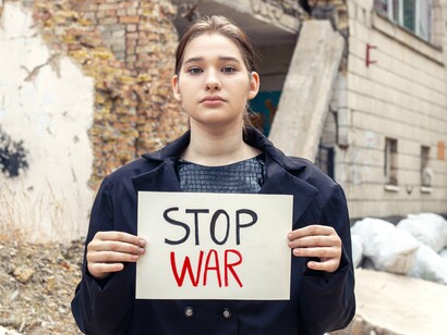 A young girl holding a “stop war” sign among ruins, representing the innocence often lost between military alliances and strategic decisions