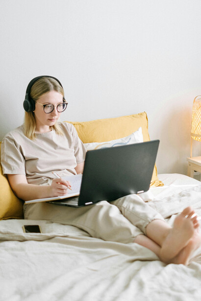 A woman sits on her bed using a black laptop, reflecting a sedentary lifestyle