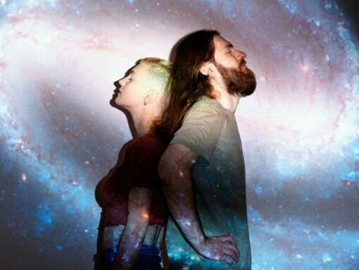 Un uomo e una donna sono in piedi schiena contro schiena, entrambi con lo sguardo rivolto verso l'alto, immersi in una proiezione di una galassia