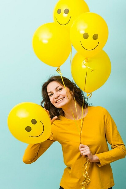 A smiling woman posing in the front view, holding balloons with smiling faces on them, symbolizing happiness