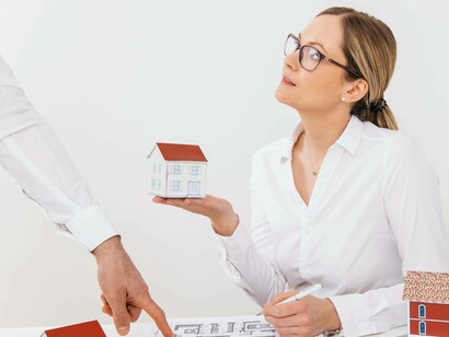 Una donna, con gli occhiali, tiene in mano un modellino di casa e guarda un collega che indica una planimetria su una scrivania. Sul tavolo sono presenti altri modellini di case