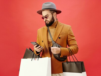 A man in a brown blazer walking with shopping bags and checking his phone, symbolizing how consumerism intersects with the search for connection and identity