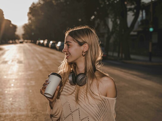 A smiling blonde woman enjoys a warm drink while listening to music or language lessons through her headphones