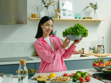 A woman in pink long sleeves stands in the kitchen holding a bowl of lettuce