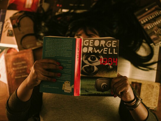 Person sitting and reading George Orwell’s 1984, surrounded by a dim, reflective atmosphere