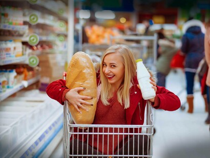 Una giovane donna all'interno di un carrello della spesa, che tiene in mano un grande pane e una bottiglia di latte