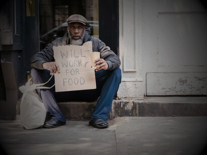A mature man sits in a doorway holding a cardboard sign, representing homelessness and poverty in the United States