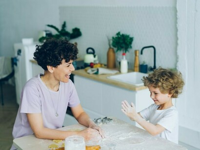A woman and a child are sitting at a table, with the child happily clapping their hands in flour while making pastry with their mom at home