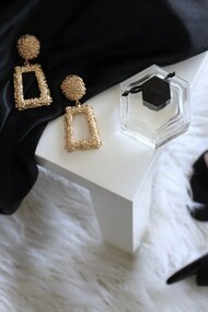 A pair of gold-colored earrings resting on a table, accompanied by black ankle-strap pumps placed on an area rug