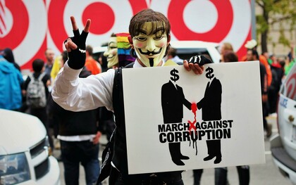 Person wearing a Guy Fawkes mask and holding a sign that reads “March Against Corruption”