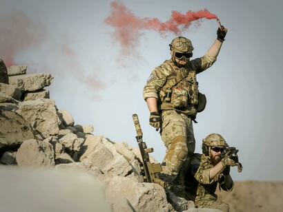 Due soldati in tenuta da combattimento sono appostati su un cumulo di macerie. Uno, in piedi, alza una torcia fumogena rossa, creando una scia di fumo. L'altro è accovacciato, con l'arma puntata in avanti