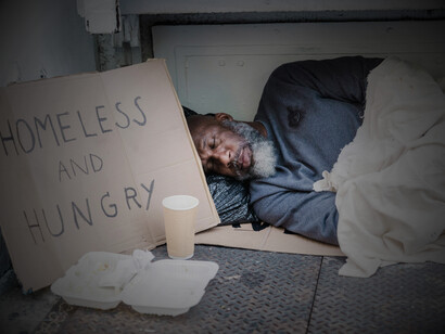 A photo of a homeless man sleeping on the ground with a cardboard sign placed beside him, in the US