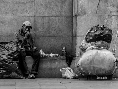 A man sits alone on a cold sidewalk, a stark reminder of how systemic inequality leaves millions in economic exile despite global wealth accumulation