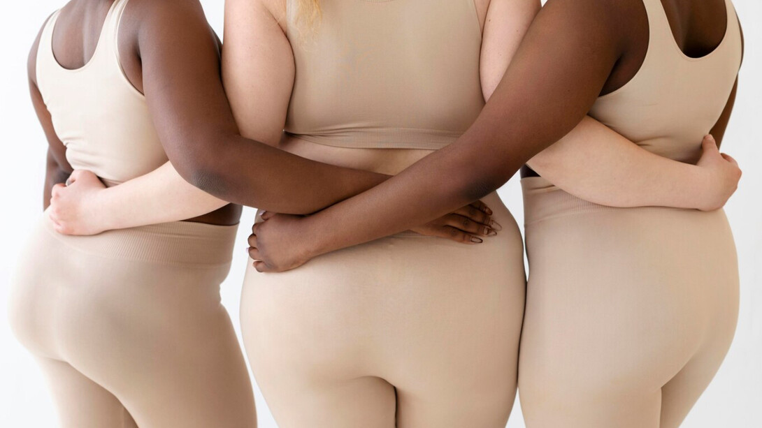 Three women stand together, arms intertwined, their bodies visible rejecting narrow ideals 