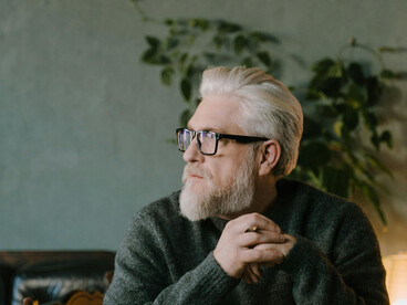 An elderly man, immersed in contemplation, writes at a table with a pen in hand