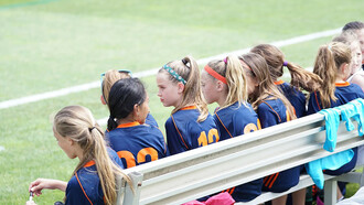 Female football players sitting on a bench during a daytime match, resting between plays