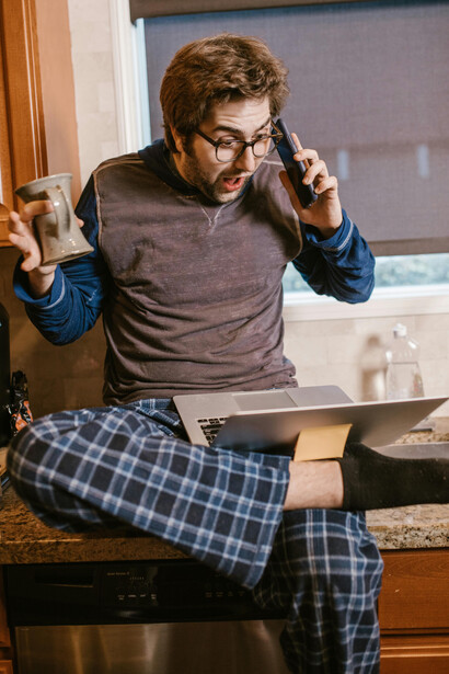 At the kitchen counter, a man excitedly juggles a phone call and the news on his computer, not realizing he's caught up in fake news
