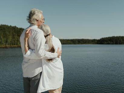 A mature couple shares a warm hug near the water’s edge, smiling with contentment