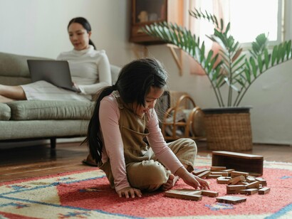 Criança brinca livremente enquanto a mãe trabalha no computador. Talvez seja necessário reaprender com as próprias crianças. Elas ainda sabem que o mundo pode ser reinventado com um pedaço de pau, um papel colorido, uma música desafinada. Elas ainda acreditam que monstros existem, que voar é possível, que o tempo pode parar numa tarde de chuva. É essa sabedoria ancestral que estamos perdendo — e, com ela, perdemos também parte essencial da nossa humanidade