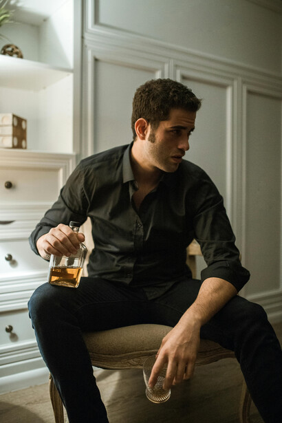 Seated on the couch, a man holds a bottle of alcohol, confronting the weight of routine