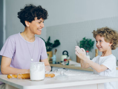 A woman and a child sit together at a table, the child excitedly clapping flour on their hands as they bake pastry with their mom at home