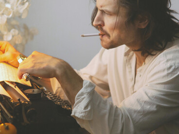 In a dimly lit room, a solitary poet sits at his desk, cigarette in hand, lost in thought over a sheet of paper