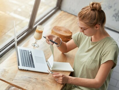 Young woman in a cozy café, wearing glasses, thoughtfully typing on her laptop with AI assistance