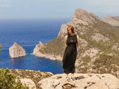 A young woman with red hair stands with her eyes closed on a rock, overlooking the mountains of the Formentor Peninsula in Mallorca