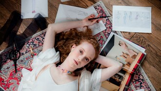 A fashion designer in a white blouse lies on a carpet, working on her sketches