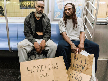 Two people sitting side by side in a subway, holding a handwritten sign that states they are homeless, representing housing insecurity in everyday public spaces