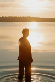 A figure stands at the shore of a lake, illuminated by the sun. 