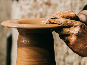 A man shapes a clay vase in his sunlit studio, with craft tools and materials spread across the desk