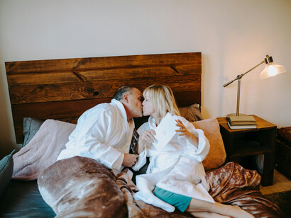 An elderly couple kisses warmly while lying on a bed in white robes, radiating happiness