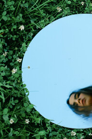 Amidst a lush green field, she gazes intently at her reflection, reminding us that in storytelling, true emotional depth comes from embracing vulnerability