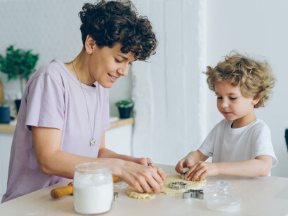 A woman and her child are seated at a table, with the child joyfully clapping flour-covered hands while baking pastry with their mom at home