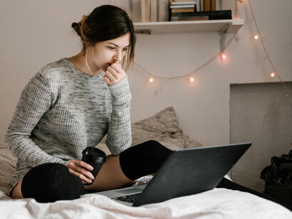 A woman in a gray sweater sits on a bed surrounded by books and a laptop in a minimalist room