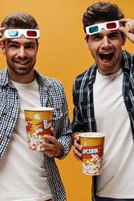 Two men in white T-shirts and blue checkered shirts, holding popcorn and removing 3D glasses, watching a movie against an isolated orange background
