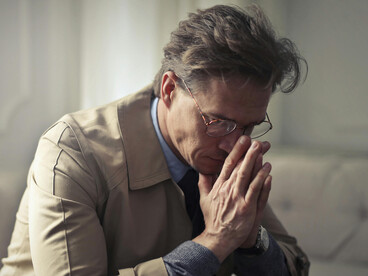 A man in a brown jacket sits on a couch, lost in thought