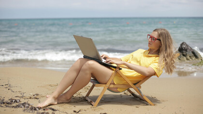 A woman in a yellow shirt sits on a wooden folding chair at the beach, working peacefully by the sea
