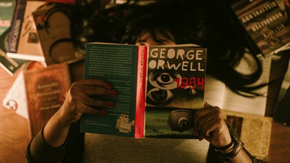 Person sitting and reading George Orwell’s 1984, surrounded by a dim, reflective atmosphere