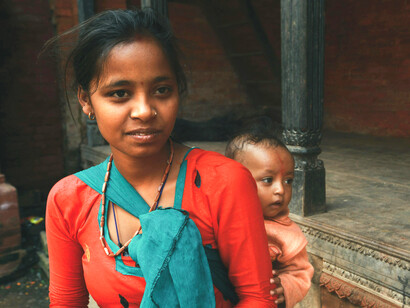 A young mother holding her baby in a traditional setting, symbolizing poverty and the barriers that keep many girls out of school