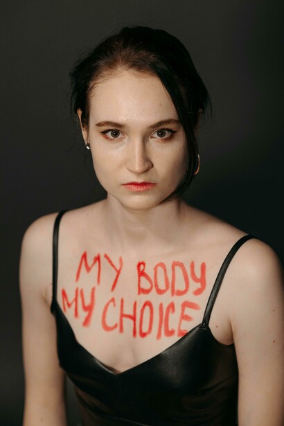 A women stares directly at the camera with the word boldly painted in red across her chest — an assertion of bodily autonomy in a world that demands explanation 