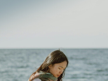 A peaceful scene of an Asian woman with long hair gazing at the ocean from the shoreline, capturing solitude and gentle loneliness