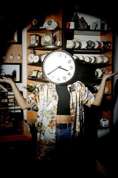 A figure stands before shelves crowded with clocks, concealing their face behind one—reflecting the way modern life submerges identity beneath the relentless pressure of time