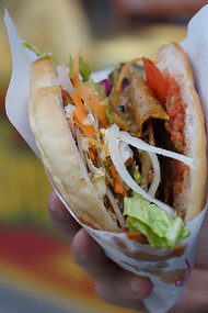 A man holding a doner kebab in front of a bustling street food restaurant