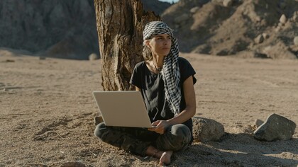 A woman in a black shirt sits on the ground in the desert, working remotely on her laptop