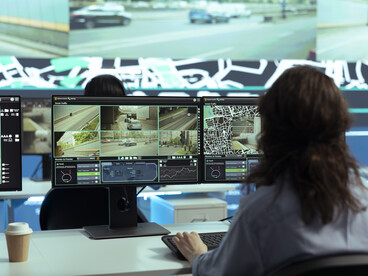 Un operatore che monitora numerosi schermi che visualizzano flussi di traffico e mappe dettagliate di navigazione GPS. Questa attività di controllo capillare e gestione centralizzata dei dati può essere vista come un'espressione della capacità delle multinazionali di praticare le proprie strategie fuori dal contesto legale, contribuendo allo Stato condominiale e al successivo Stato della sorveglianza