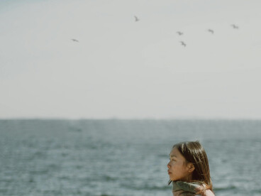 An Asian woman with long hair standing by the sea, calmly observing the waves, evoking a quiet sense of solitude and loneliness
