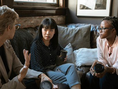 A group of diverse women, including a Black woman, an Asian woman and a Caucasian woman, talk at work while holding disposable cups