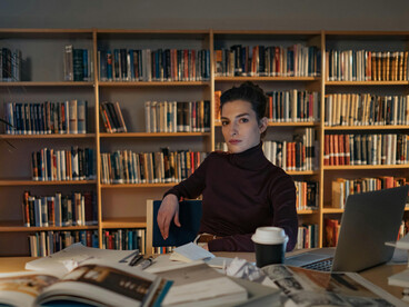 A brunette woman works alone in a home office late at night, lit only by her computer screen